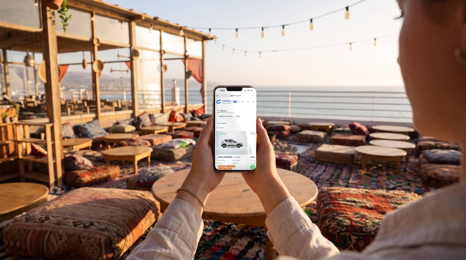 Voyageuse consultant les voitures GadiWaze disponibles à Taghazout sur son téléphone, dans un café rooftop face à l'océan