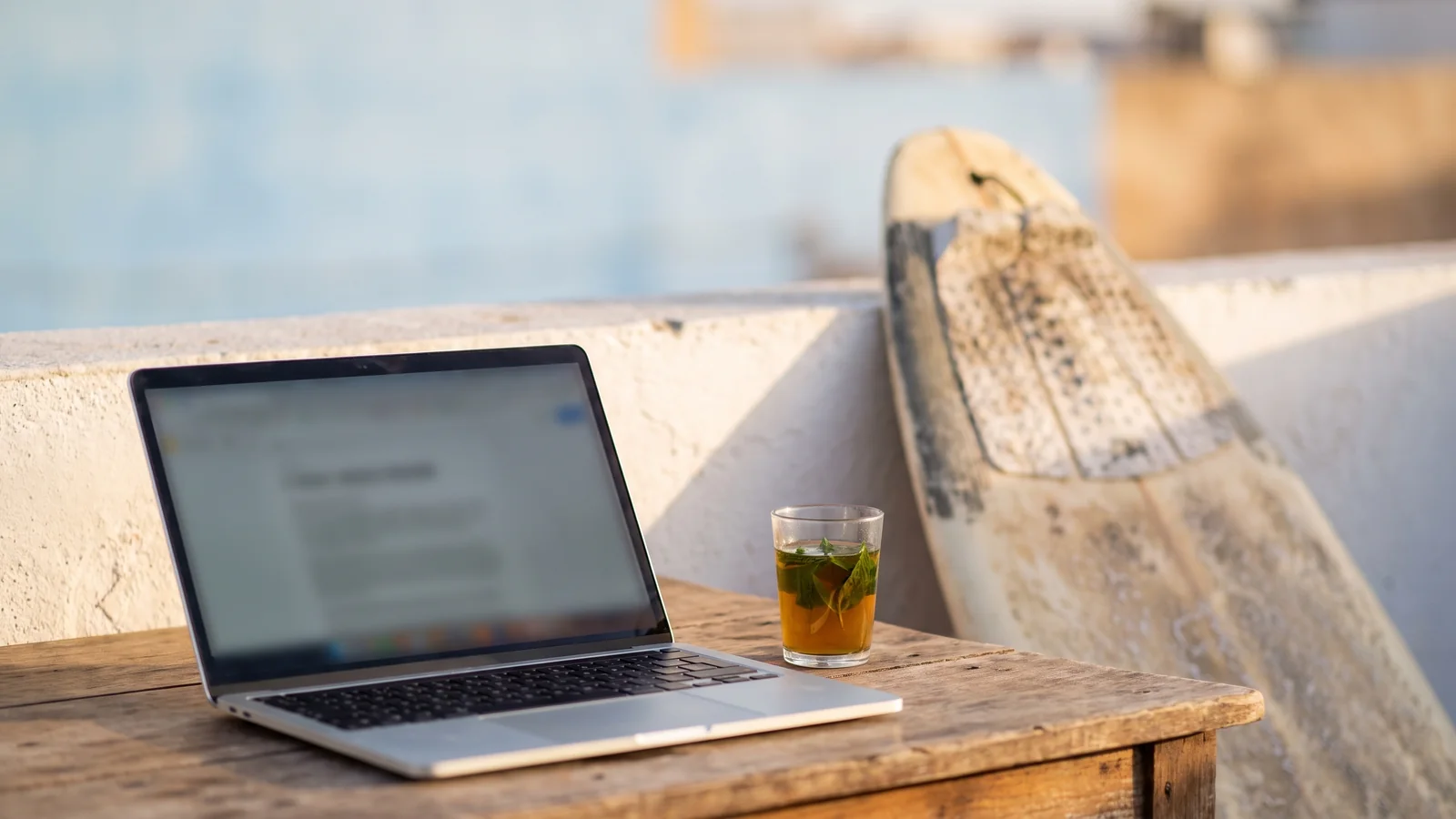 Laptop and mint tea on a rooftop terrace, a surfboard leaning behind, the ocean blurred in the distance